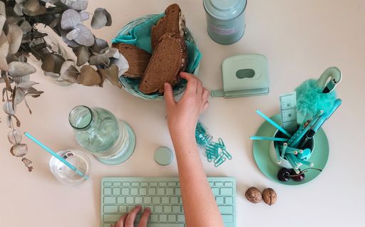 Different turquoise colored objects like computer keyboard, pens and paper clips; and hand grabbing a piece of bread