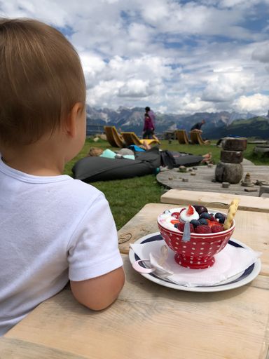 18 months old toddler sitting at a wooden table with red bowl of cereals next to it looking out on a scenic view on Seiser Alm and its surroundings