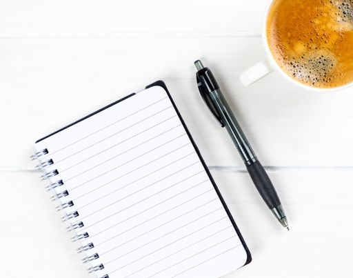 A closeup of a coffee cup with paper and pen on a white wooden background