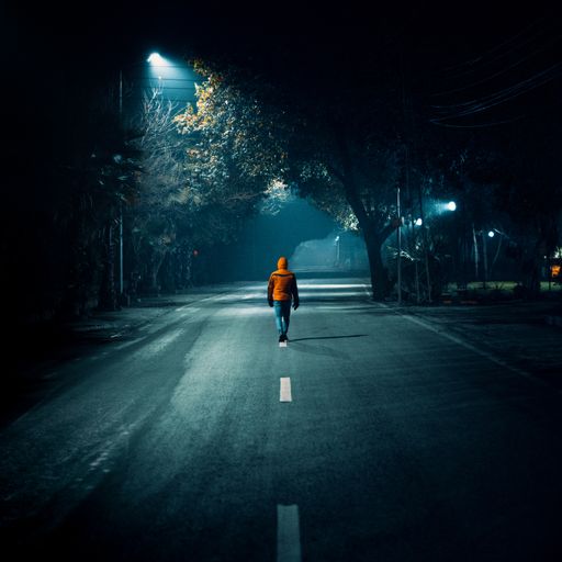 A beautiful shot of a young man wearing an orange jacket walking in the middle of a street with a street light in the night