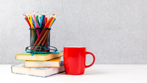 A closeup of books, colorful pencils, glasses and red cup on a desk with gray background