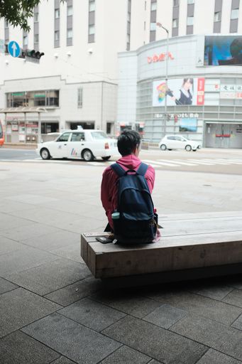 A vertical selective shot of a person wearing black backpack sitting on a bench  on the sidewalk near cars and buildings