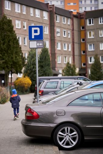 A row of parked cars on parking places with sign by a sidewalk with child on a scooter in the Stare Zegrze district.
