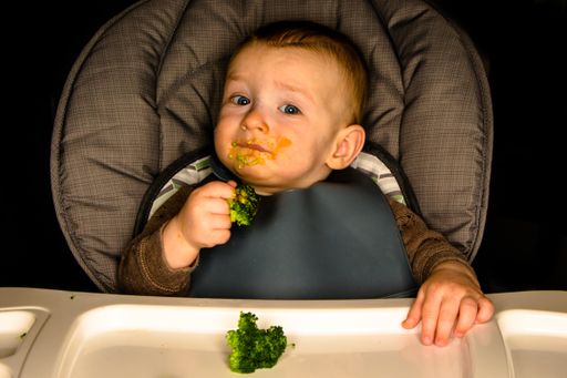 A closeup of a male baby sitting in a high chair holding broccoli with brown sauce and bits all over his mouth on a black background