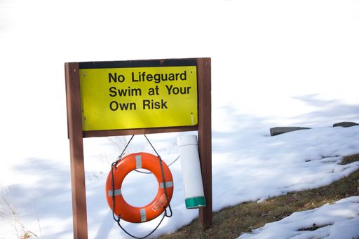 A wooden warning yellow sign with "No Lifeguard, Swim at Your Own Risk" text in black with hanging life-ring in a snowy field on a sunny day