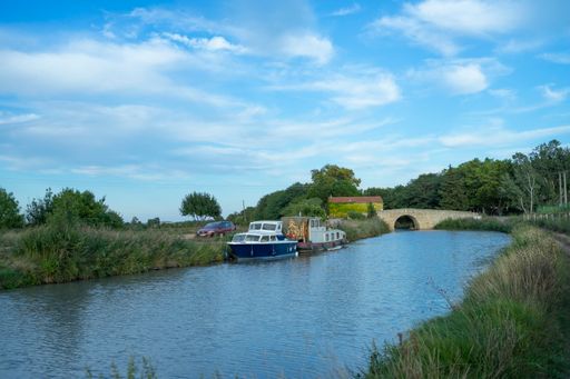 A scenic shot of the Canal Du Midi sewer river with a bridge and boats under a cloudy blue sky