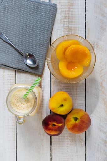 A top view of fresh ripe peaches, natural peach juice or nectar, and canned peach halves in a glass bowl on a wooden table