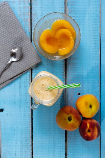 A top view of fresh ripe peaches, natural peach juice in a mason jar, and canned peaches in a glass bowl on a blue wooden background