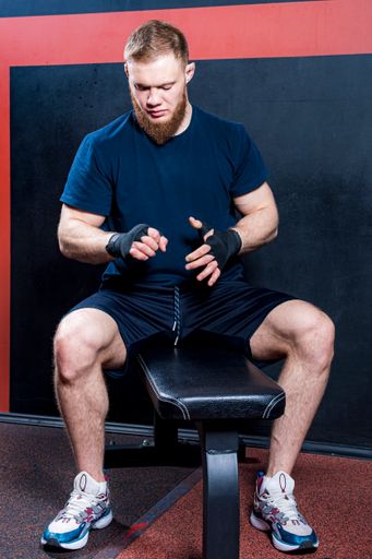 Young bearded mixed martial arts fighter sits on sports bench and examines his hands, which are wrapped in protective elastic bandage.