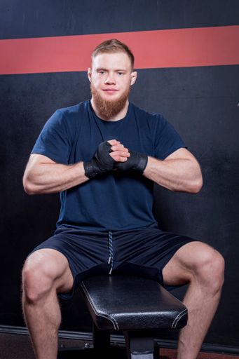 Young bearded mixed martial arts fighter sits on sports bench, clenched his fists in elastic bandages in front of his chest and looks at camera.
