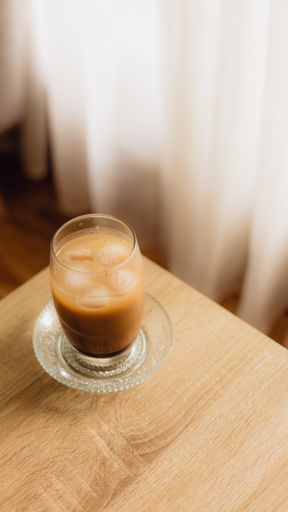 A top view shot of an Iced coffee glass cup on a wooden table with curtains in the background at home