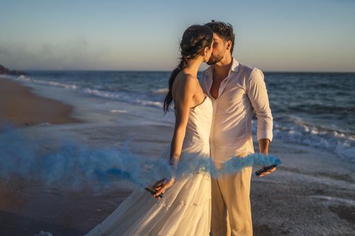 A caucasian loving couple holding a blue colored smoke and kissing in the beach during a wedding photoshoot