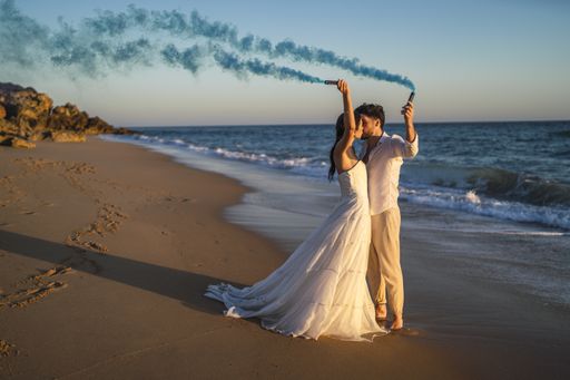 A caucasian loving couple holding a blue colored smoke and kissing in the beach during a wedding photoshoot