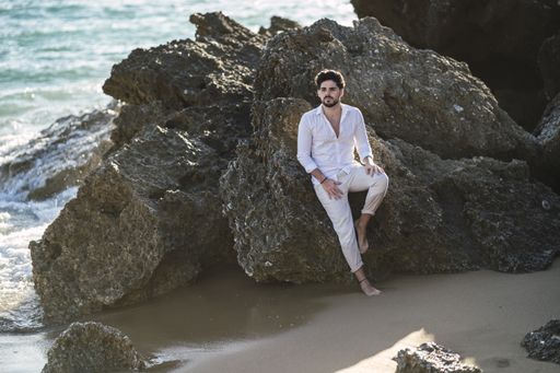 A caucasian man wearing white clothes sitting on the stone on the beach
