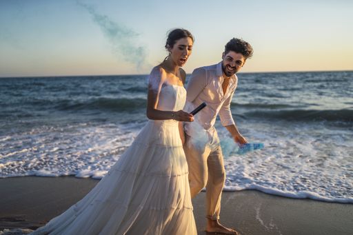 A caucasian loving couple holding a blue colored smoke with a scarred face in the beach during a wedding photoshoot