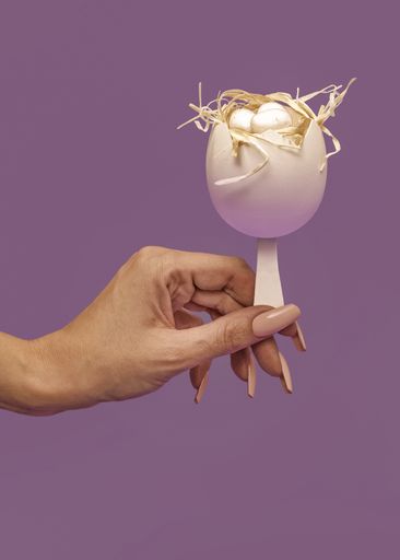 A female hand holding a decorative white egg on an ice cream stick on isolated purple background