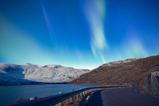 A horizontal shot of an empty road near the lake surrounded by high mountains covered in snow under the cloudy sky
