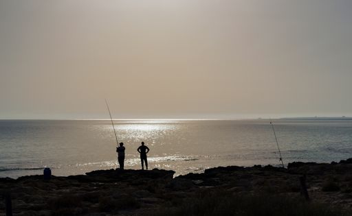 A silhouette of two person standing on the sea and fishing during sunset