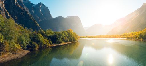 A horizontal shot of a beautiful lake surrounded by green trees and high mountains during the sunrise