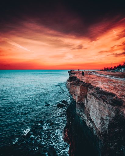 A vertical shot of a man on the edge of a cliff near the sea with turqoise blue water and red sunset