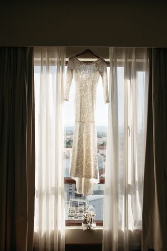 A vertical of a female white wedding dress hanging on a window with high-heeled shoes on windowsill