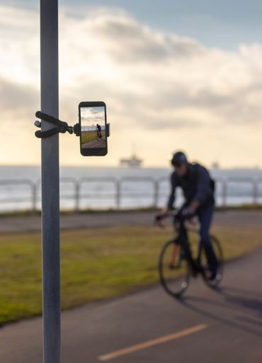 A selective focus of a tripod holding a phone on metallic post shooting a male riding bicycle on the Huntington beach, California
