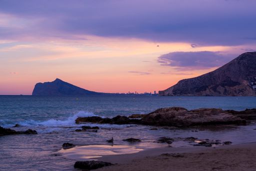 The wavy ocean hitting the rocky beach surrounded by mountains under the pink cloudy sky