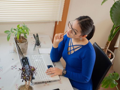A young smiling Asian girl with glasses and headphones working from home with a laptop and cell phone