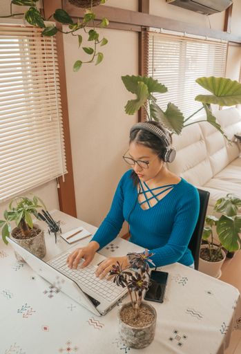 The Asian girl with glasses, headphones and plants working from home with a laptop and cell phone.