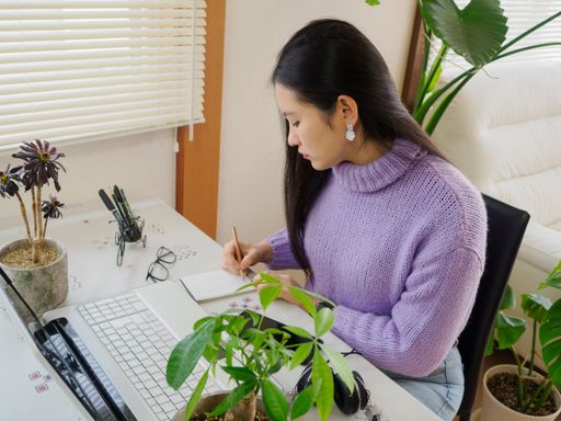 A young Asian girl working from home at her desk with a laptop and headphones surrounded by plants