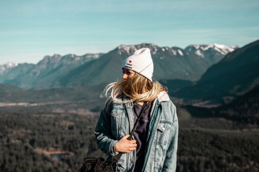 A selective shot of a woman wearing a white hat and jean jacket standing on a cliff with a beautiful view behind her