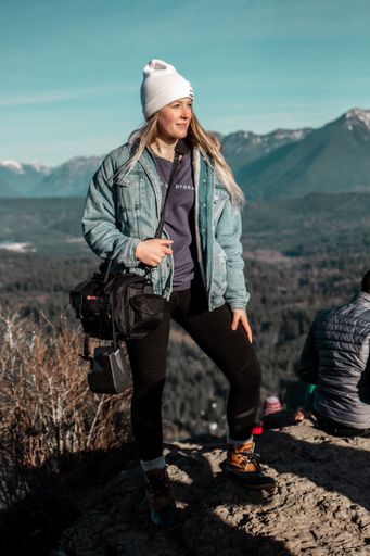 A selective focus shot of a woman wearing a white hat and jean jacket standing on a cliff with a beautiful view behind her