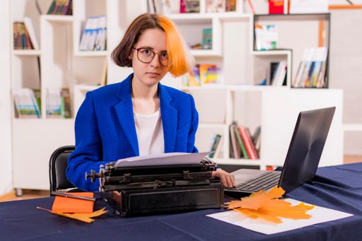 Young girl with multi-colored hair and glasses sits at office desk. In front of her are old typewriter and modern laptop. Looks into camera.
