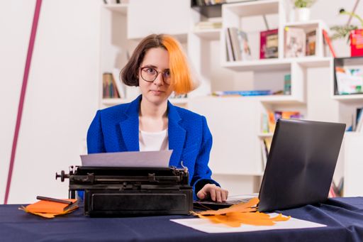 Young girl with multi-colored hair and glasses sits at office desk. In front of her are old typewriter and modern laptop. Looks into camera.