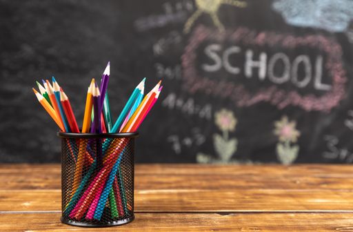 A closeup shot of colored pencils in mesh pencils holder on wooden table with chalkboard school in the blurred background