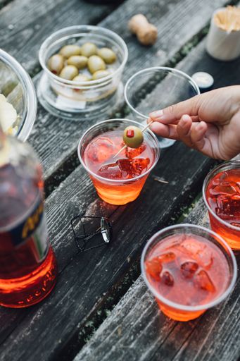 Woman hand placing green olive into Aperol spritz sparkling aperitif cocktails outdoors on picnic table