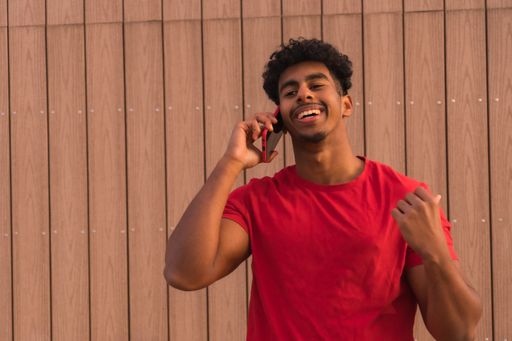 A Middle Eastern young man in a red t-shirt in front of a wooden background talking on the phone