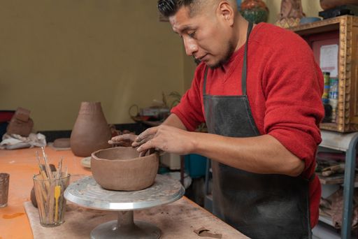 A side shot of a Hispanic potter making and molding a clay vessel with both hands in his workshop