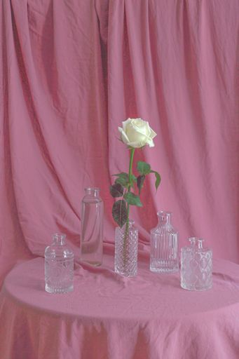 A vertical shot of white roses with beautiful glass bottles on a pink fabric surface and background
