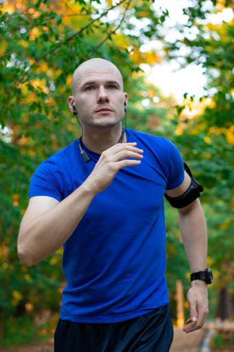 A bald male wearing a blue shirt exercising and running in nature with green trees all around