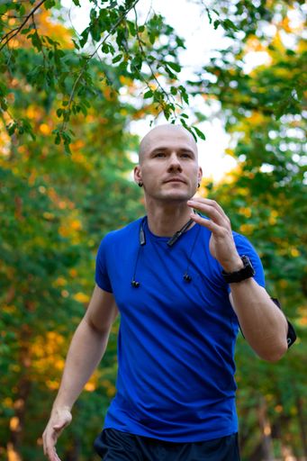 A bald male wearing a blue shirt exercising and running in nature with green trees all around