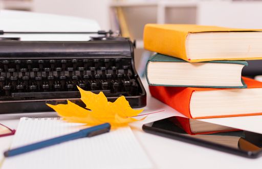 Autumn mood. On table are old typewriter, modern mobile phone, books, notebook and pen, dry yellow maple leaves. Workplace of student or writer.