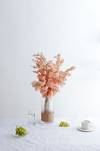 The vertical still life shot of decorative flowers on a vase with a teacup and a glass and grapes on a white table