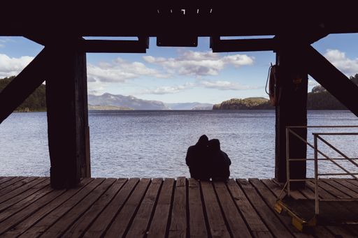 A back shot silhouetted couple sitting on wooden boards floor under a bridge looking at the water sea