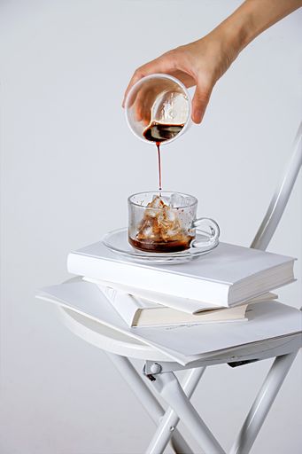 A vertical shot of a hand gradually adding coffee to a cup with ice cubes on a white background