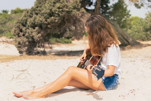 A young caucasian woman playing guitar on the beach with trees in the background.