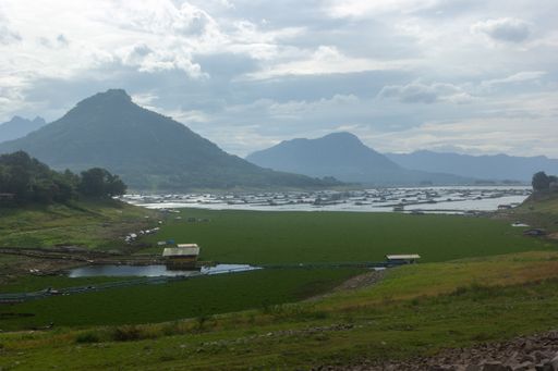 A scenic view of a green field with old houses and lakes against mountains in clouds in Jatiluhur Reservoir, Indonesia