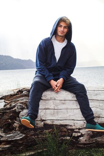 A bright vertical shot of a Caucasian male wearing a hoodie, sitting on a wooden log in front of lake