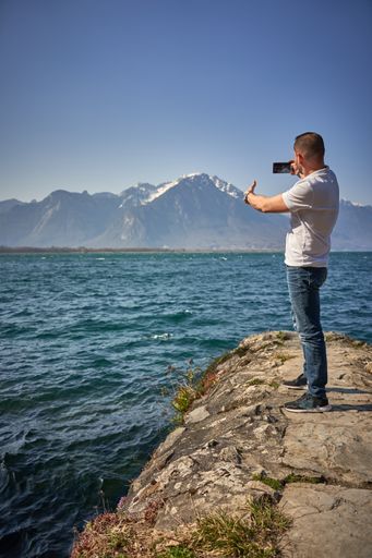 An adult man standing on rocks at the lakeshore taking a selfie against alps mountains on a sunny day in Switzerland