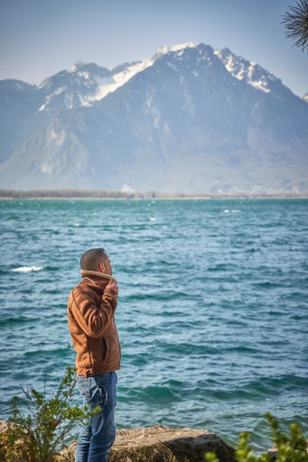An adult man standing on rocks at the lakeshore against alps mountains on a sunny day in Switzerland